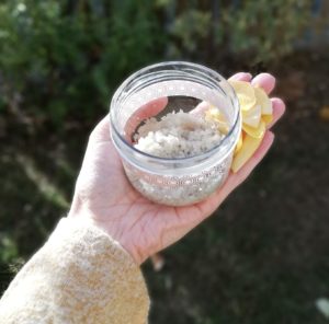 Image d'un pot du mélange pour le gommage dans une main accompagnée de pétales de fleur jaune